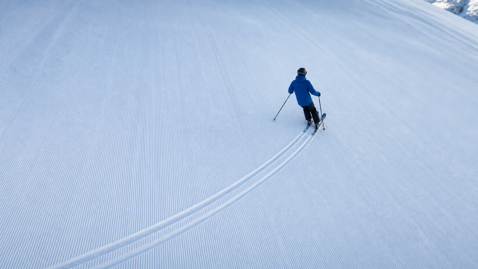 A skier on a groomed slope makes a broad turn, leaving two parallel tracks
